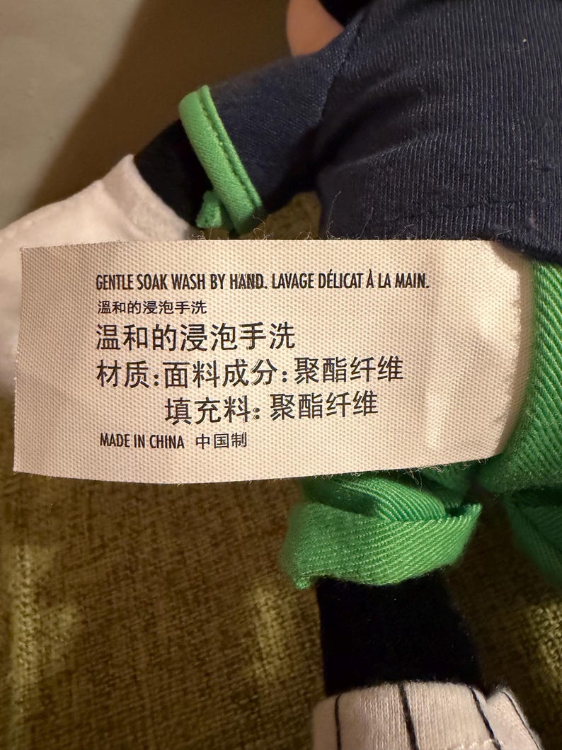 May include: A plush toy with a tag that reads "GENTLE SOAK WASH BY HAND" in English and French. The tag also includes Chinese characters. The toy has a navy blue and green outfit with white and black accents.