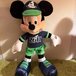 May include: A Mickey Mouse plush toy wearing a navy blue cap and shirt with the year "2017" printed on the shirt. The plush has green shorts and shoes with blue and green stripes. The toy has black ears and white gloves.