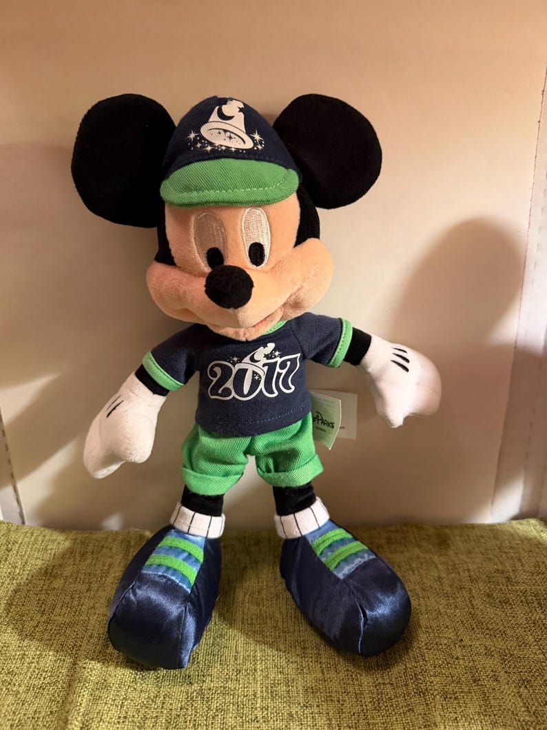 May include: A Mickey Mouse plush toy wearing a navy blue cap and shirt with the year "2017" printed on the shirt. The plush has green shorts and shoes with blue and green stripes. The toy has black ears and white gloves.