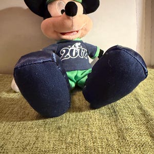 May include: A plush Mickey Mouse doll wearing a navy blue shirt with the number "2012" printed on it, green shorts, and a green and navy blue baseball cap. The doll has large black ears and blue feet.