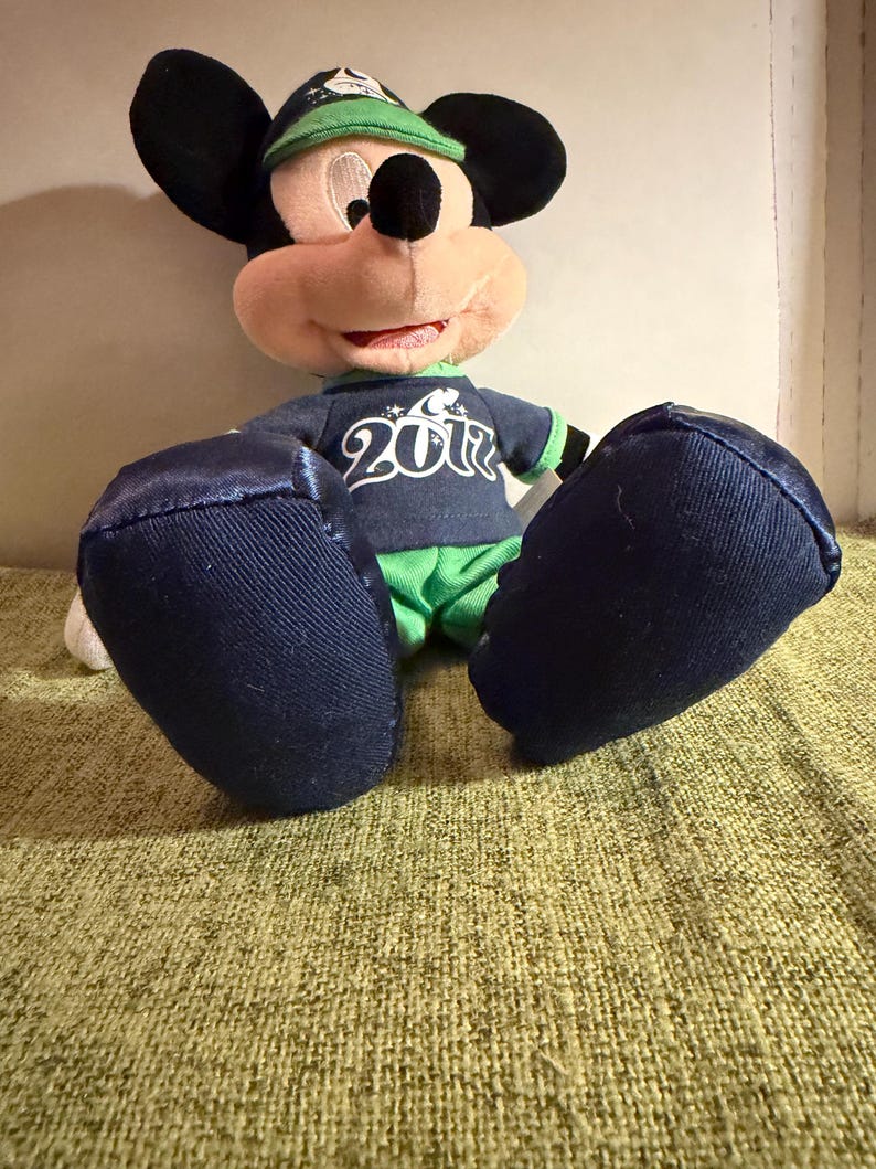 May include: A plush Mickey Mouse doll wearing a navy blue shirt with the number "2012" printed on it, green shorts, and a green and navy blue baseball cap. The doll has large black ears and blue feet.