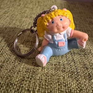 May include: A keychain featuring a small figurine of a child with blonde hair in pigtails, wearing a white shirt and light blue overalls with pink accents. The figure has blue eyes and is attached to a silver metal split ring by a short chain.
