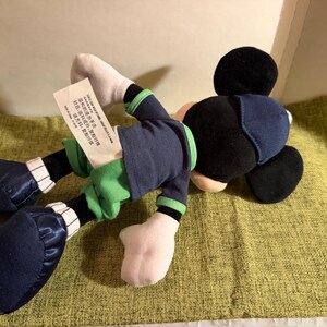 May include: A plush Mickey Mouse doll, lying on its side. The doll features a black head and ears, a navy blue outfit with green accents, white gloves, and navy blue shoes. A tag is attached to the doll.