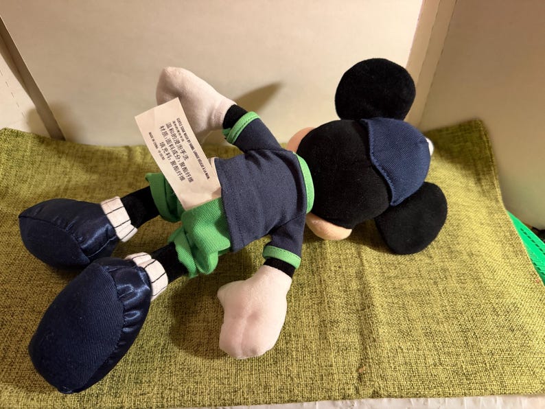 May include: A plush Mickey Mouse doll, lying on its side. The doll features a black head and ears, a navy blue outfit with green accents, white gloves, and navy blue shoes. A tag is attached to the doll.