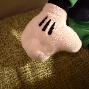 May include: Close-up of a plush toy hand, featuring a white glove with three black lines representing fingers. The hand is attached to a dark blue sleeve with a green cuff. The toy is resting on a textured green surface.