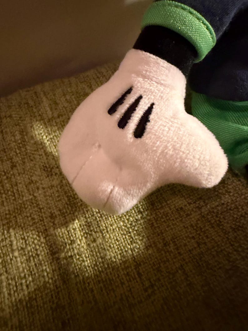 May include: Close-up of a plush toy hand, featuring a white glove with three black lines representing fingers. The hand is attached to a dark blue sleeve with a green cuff. The toy is resting on a textured green surface.