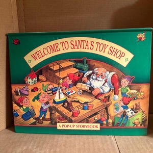 May include: A green pop-up storybook titled "Welcome to Santa's Toy Shop" with illustrations of Santa Claus and elves making toys in a workshop.
