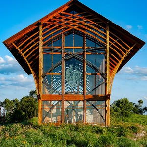 Op de afbeelding: Een houten kapel met een groot glazen raam met een glas-in-loodontwerp. De kapel is omgeven door groen gras en een blauwe lucht.