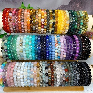 Op de afbeelding: Een display van kleurrijke kralenarmbanden gemaakt van verschillende edelstenen, waaronder amethist, turquoise en tijgeroog. De armbanden zijn gerangschikt in rijen op een houten standaard met gouden accenten.