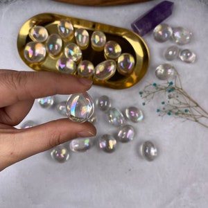 May include: Iridescent crystal stones in various shapes and sizes. Some are in a gold-coloured tray, others scattered on a white surface. One stone is held between fingers, showing its rainbow sheen.