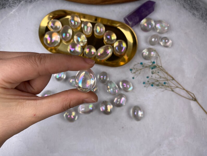 May include: Iridescent crystal stones in various shapes and sizes. Some are in a gold-coloured tray, others scattered on a white surface. One stone is held between fingers, showing its rainbow sheen.