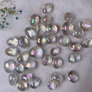 May include: A scattering of clear, iridescent quartz stones, each with a polished, rounded form. The stones display a spectrum of colours, arranged on a textured, off-white surface. Delicate dried flowers with small blue blooms are in the upper left corner.