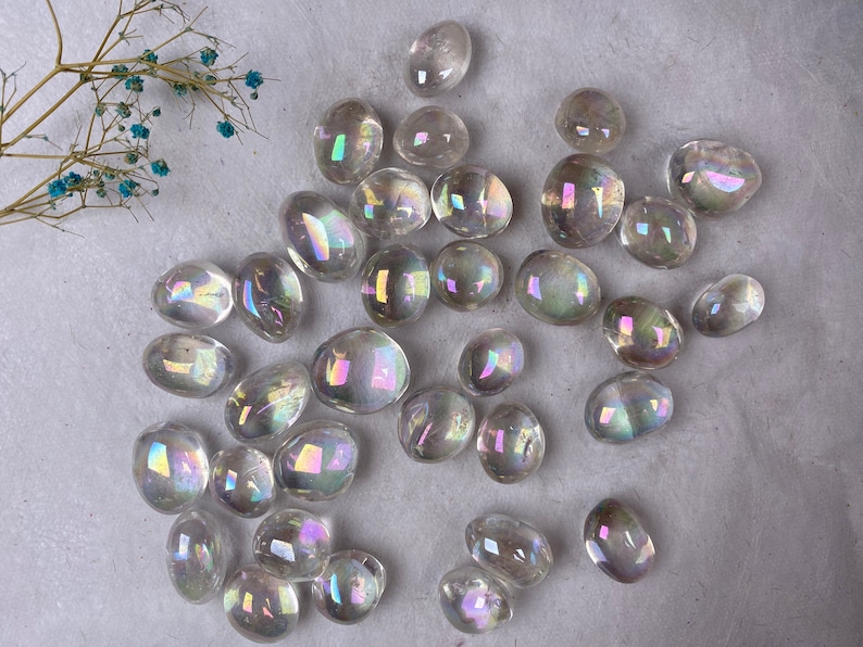 May include: A scattering of clear, iridescent quartz stones, each with a polished, rounded form. The stones display a spectrum of colours, arranged on a textured, off-white surface. Delicate dried flowers with small blue blooms are in the upper left corner.