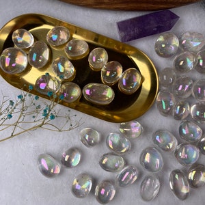 May include: Iridescent polished stones with a rainbow sheen are arranged on a gold tray and scattered on a white surface. A purple crystal and dried flowers are also present. The stones vary in size and shape.