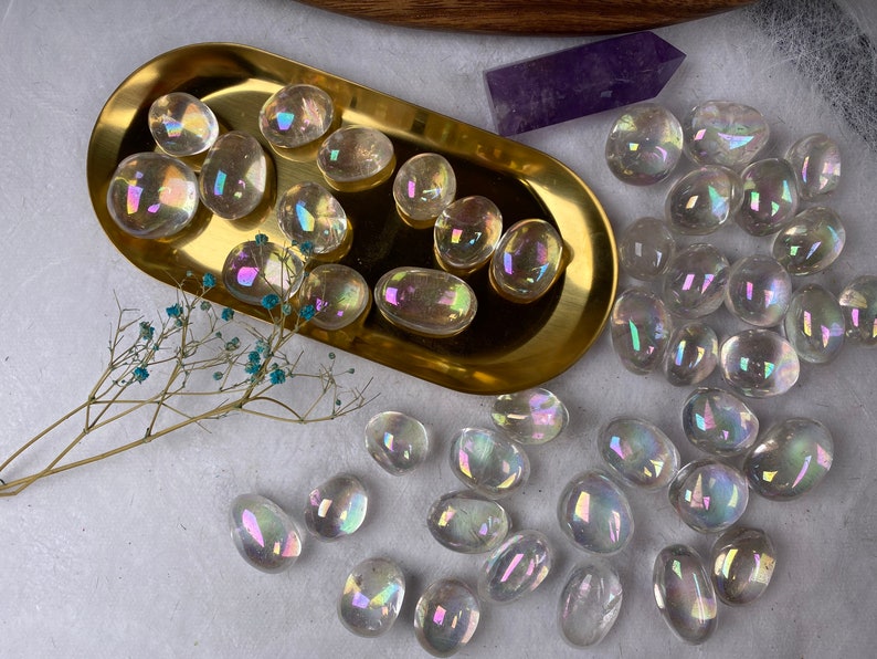 May include: Iridescent polished stones with a rainbow sheen are arranged on a gold tray and scattered on a white surface. A purple crystal and dried flowers are also present. The stones vary in size and shape.