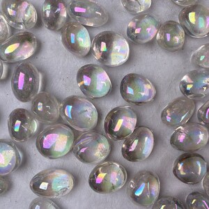 May include: A selection of clear, polished stones with an iridescent, rainbow-like sheen. The stones are oval and teardrop-shaped, reflecting light with hues of pink, yellow, and blue. They are scattered across a neutral surface.