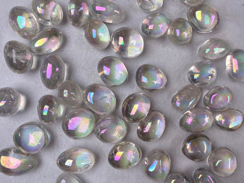 May include: A selection of clear, polished stones with an iridescent, rainbow-like sheen. The stones are oval and teardrop-shaped, reflecting light with hues of pink, yellow, and blue. They are scattered across a neutral surface.