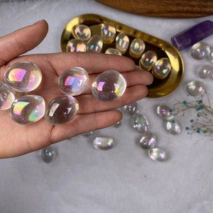 May include: A hand holding several clear, iridescent aura quartz stones. The stones have an oval shape and reflect a rainbow of colours. Other stones are in a gold tray and scattered on a white surface. A purple crystal and dried flowers are also present.