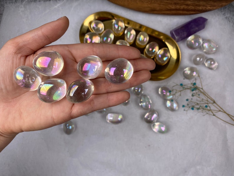May include: A hand holding several clear, iridescent aura quartz stones. The stones have an oval shape and reflect a rainbow of colours. Other stones are in a gold tray and scattered on a white surface. A purple crystal and dried flowers are also present.