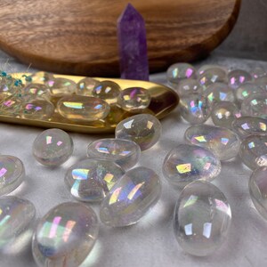 May include: A selection of clear, iridescent aura quartz stones. The stones are oval-shaped and reflect a rainbow of colours. A purple crystal point and a gold-coloured tray with small stones are in the background.
