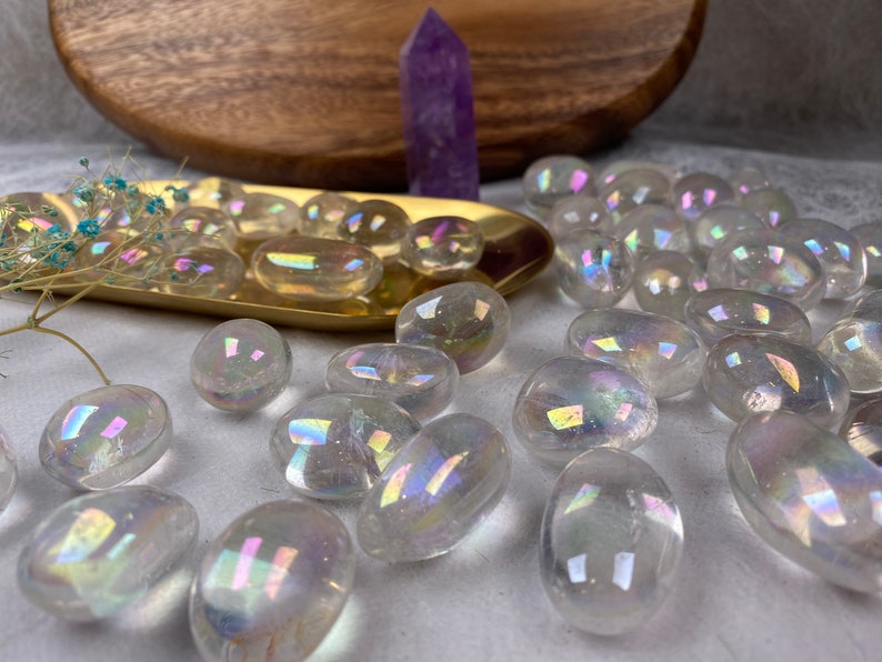 May include: A selection of clear, iridescent aura quartz stones. The stones are oval-shaped and reflect a rainbow of colours. A purple crystal point and a gold-coloured tray with small stones are in the background.