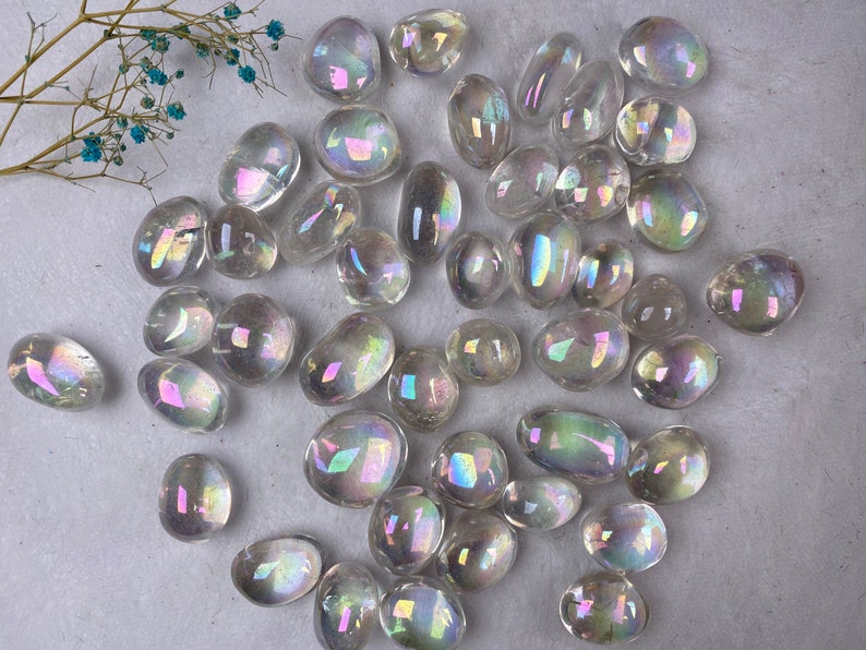 May include: A selection of clear, iridescent tumbled stones with a rainbow sheen. The stones are of various sizes and shapes, with a smooth, polished texture. A sprig of dried blue flowers is in the upper left corner.