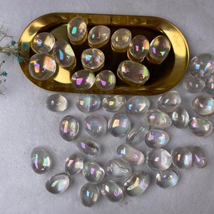 May include: A selection of iridescent quartz stones, clear with a rainbow sheen, in a gold-coloured oval tray. The stones vary in size and shape. A sprig of dried flowers with small blue flowers is in the top left corner.
