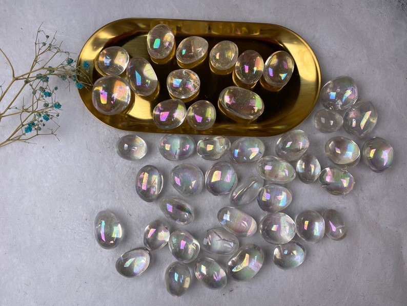 May include: A selection of iridescent quartz stones, clear with a rainbow sheen, in a gold-coloured oval tray. The stones vary in size and shape. A sprig of dried flowers with small blue flowers is in the top left corner.