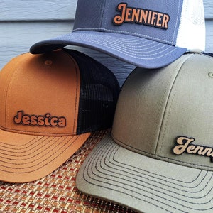 May include: Three trucker hats with black mesh backs. The hats are brown, gray, and green. The hats have custom names on them: Jessica, Jennifer, and Jennifer.