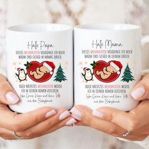 Op de afbeelding: Twee witte keramische mokken met kerstillustraties. Eén mok zegt "Hallo Papa" en de andere "Hallo Mama". Elke mok heeft een baby in een hart, een pinguïn en een kerstboom. De mokken worden vastgehouden.