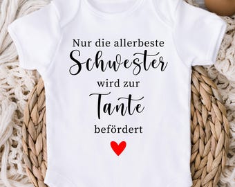 May include: A white short-sleeved baby bodysuit featuring the black text "Nur die allerbeste Schwester wird zur Tante befördert" and a small red heart icon centered below the text. The garment has a round neckline and snap closures at the bottom.