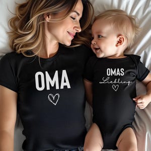May include: Black t-shirt with the word "OMA" and a heart graphic. A baby is wearing a black onesie with the words "OMAS Liebling" and a heart. The word "SCHWARZ" is at the bottom. Both are lying on a white bed.