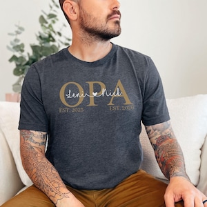 Könnte beinhalten: Ein dunkelgrau meliertes Kurzarm-T-Shirt mit dem goldenen Schriftzug OPA und den Namen Leni und Nick in weißer Schreibschrift mit einem kleinen Herz dazwischen. Darunter steht in Gold gedruckt EST. 2023 und EST. 2026.