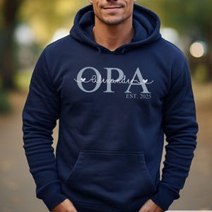 Könnte beinhalten: Marineblauer Kapuzenpullover mit Vordertasche. Das Wort "OPA" ist in Hellgrau gedruckt, darunter der Name "Alexandra" und darunter "EST. 2025". In der unteren rechten Ecke des Sweatshirts steht das Wort "MARINEBLAU".
