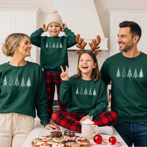 Op de afbeelding: Een gezin van vier personen draagt bijpassende donkergroene sweatshirts met witte kerstboomontwerpen. Het gezin is verzameld rond een tafel met koekjes en vakantiedecoraties. De tekst "DUNKELGRÜN" is zichtbaar.