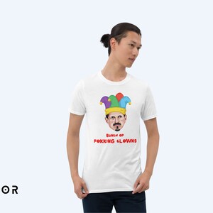 以下が含まれることがあります： 帽子をかぶった人の漫画の画像がプリントされた白いTシャツ。画像の下に「Bunch of FOKKING CLOWNS」というテキストがプリントされています。