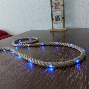 Könnte beinhalten: Ein dekoratives Seillicht mit blauen LED-Leuchten. Das Seil ist beige und liegt auf einer dunklen Holzoberfläche. Im Hintergrund befindet sich ein kleiner Holzrahmen mit drei quadratischen Öffnungen.
