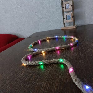 May include: A decorative rope light with multicolored LED lights. The rope is made of a thick, textured material and is arranged in a curved shape on a dark wooden surface. A wooden picture frame with three small frames is in the background.