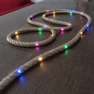 以下が含まれることがあります： マルチカラーLEDライトを備えた装飾ロープライト。ロープは自然なベージュ色で、濃い茶色の木製の表面に巻かれています。ライトは青、緑、ピンク、黄色です。