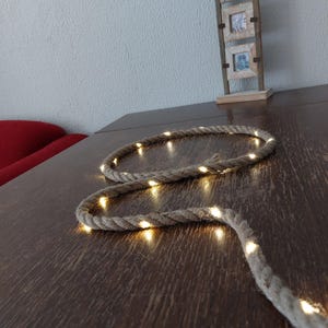 Puede incluir: Una cadena de luces LED blancas cálidas envuelta alrededor de una cuerda de yute natural. Las luces están enhebradas a lo largo de la cuerda y crean un brillo cálido.
