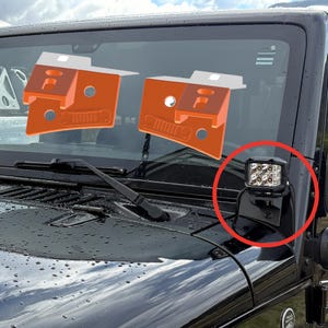 May include: A black Jeep with a light bar mounted on the front. The light bar has four square LED lights. Two orange brackets are also visible, designed to mount on the windshield frame.