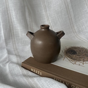 May include: A small, brown ceramic vase with two handles. The vase sits on a book titled "Nests" by Susan Ogilvy, next to a small bird's nest. The background is a white, textured fabric.