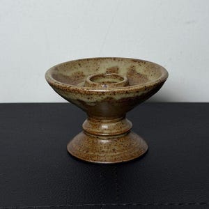 Può includere: Un portacandele in ceramica marrone con una ciotola ampia e poco profonda e una coppa centrale rialzata per la candela. Il portacandele ha una base a piedistallo e una finitura macchiata e smaltata. Il design è semplice e funzionale.