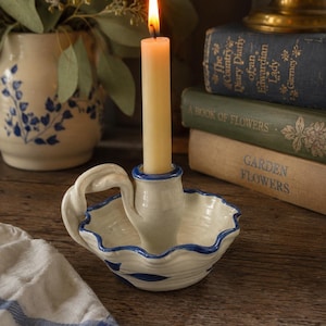 May include: A cream-colored ceramic candle holder with a scalloped edge and a blue painted rim. The holder features a looped handle and a central socket containing a lit, pale yellow taper candle. The base is decorated with blue leaf motifs.
