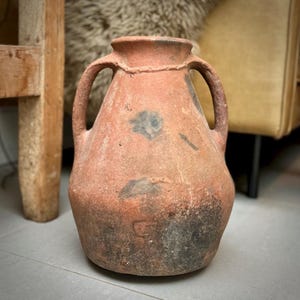 May include: A terracotta clay jar with two handles. The jar has a worn, rustic appearance with dark brown and black markings.
