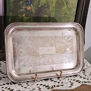 May include: A rectangular silver-toned metal tray featuring an intricate engraved floral and scrollwork pattern across the center surface. The tray has a smooth, raised border and a polished finish with visible signs of aging and patina.