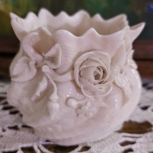 May include: A small, off-white ceramic vessel with a ruffled, scalloped rim. The exterior is decorated with raised, three-dimensional floral motifs, including a prominent rose, leaves, and small buds, all in a matching monochromatic off-white finish.