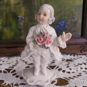 May include: A white porcelain figurine of a person dressed in 18th-century style clothing, featuring a ruffled cravat and cuffs. The figure holds a bouquet of pink roses with green leaves and stands on a scalloped base with a glossy finish.