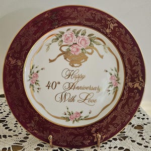 May include: A round decorative plate with a deep burgundy border featuring gold filigree patterns of leaves and birds. The center is white with a floral arrangement of pink roses, gold ribbons, and the text Happy 40th Anniversary With Love.
