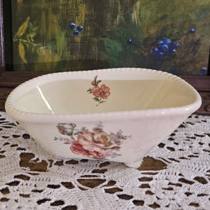 May include: A cream-colored ceramic bowl with a scalloped, rope-textured rim and four small feet. The exterior features a floral decal with pink, red, and green tones, while a single small flower decal is centered on the interior wall.